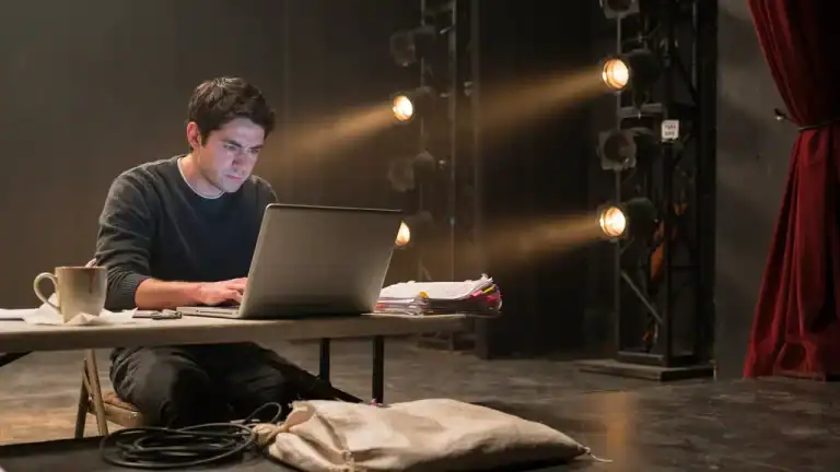 Confident actor sitting at a laptop with an AI interface on screen and stage lights in the background