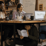 A diverse group of young professionals sits around a table in a casual office meeting, while one person discreetly reviews a script under the desk. The scene is lit with soft natural light and has a warm, candid, documentary-style feel with a touch of humor.