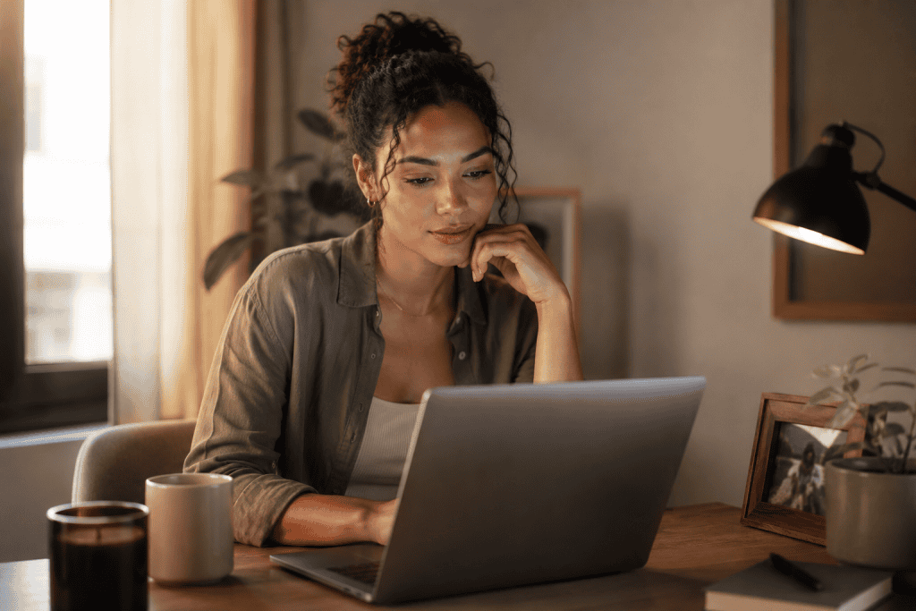 Photorealistic medium close-up of a young woman seated at a home desk, looking thoughtfully at her laptop in warm afternoon light, with a plant and framed photo nearby.